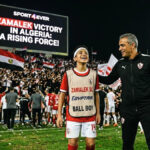A young ball boy smiling next to a coach in a stadium with Zamalek fans celebrating in the background and a large screen showing "Zamalek Victory in Algeria".