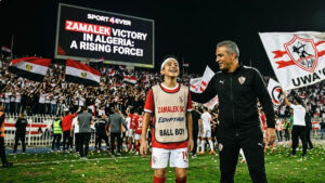 A young ball boy smiling next to a coach in a stadium with Zamalek fans celebrating in the background and a large screen showing "Zamalek Victory in Algeria".